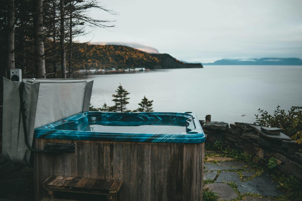A scenic view of a rustic hot tub by a calm lake surrounded by forest in overcast weather.